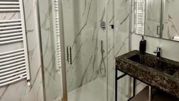 A modern bathroom with a glass-enclosed shower, a marble-tiled wall, a sink with a marble countertop, and a mirror above the sink.