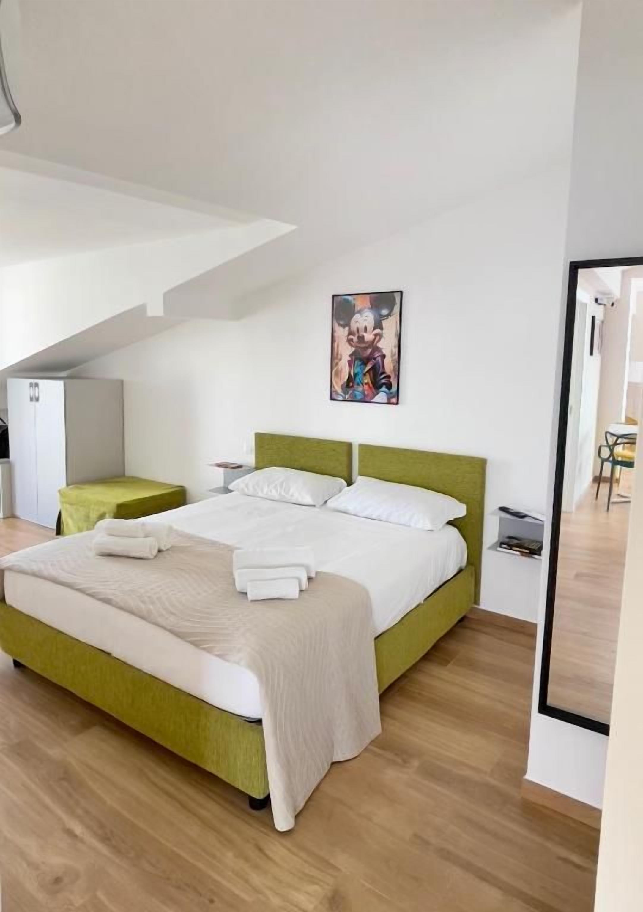 A modern bedroom with a green bed, white linens, a green nightstand, and a painting on the wall.