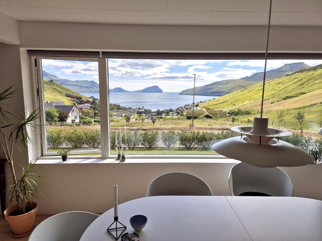 A room with a large window overlooking a scenic landscape, a dining table, and a hanging light fixture.