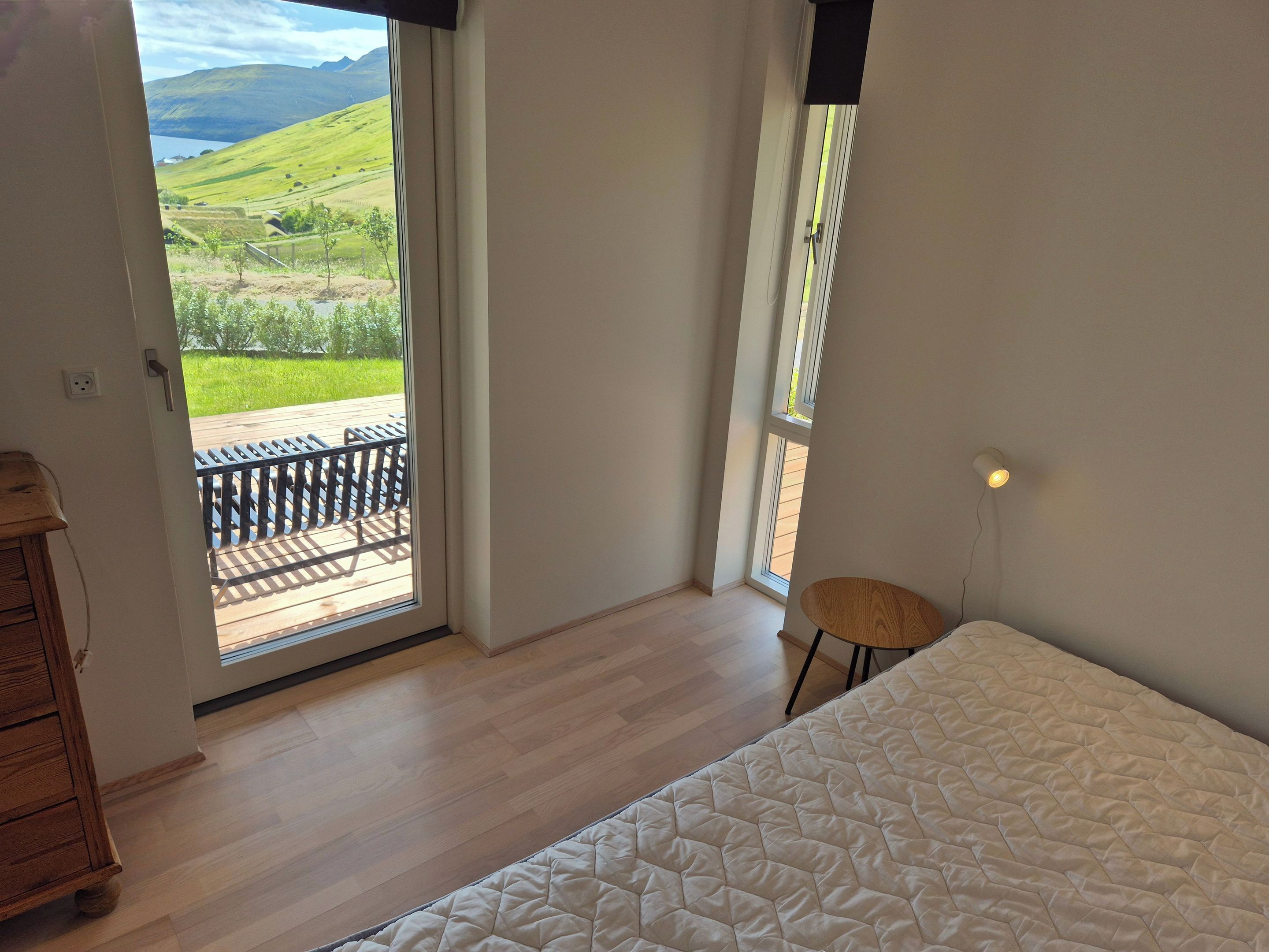 A bedroom with a wooden floor, a bed with a quilted cover, a nightstand, a chair, and a view of a green landscape through a door.