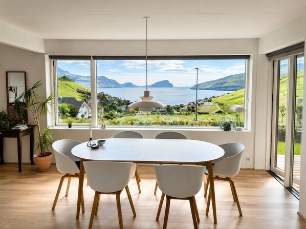A dining area with a large table and chairs, a view of a coastal landscape, and a potted plant.