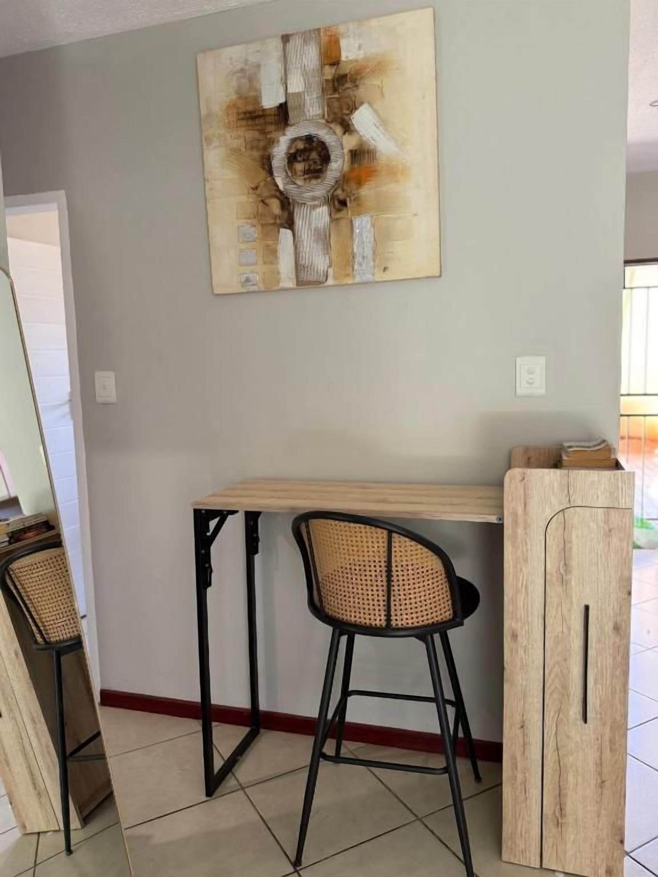 A dining area with a wooden table and two black chairs, a wall-mounted abstract painting, and a wooden cabinet.