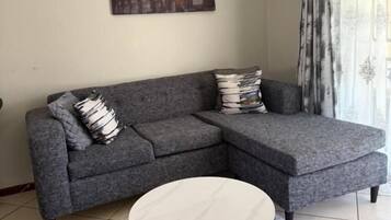 Un salon avec un canapé d’angle gris, une table basse ronde, un tapis à motifs et un tableau représentant le Colisée.