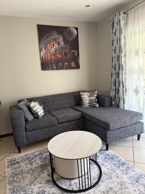 Un saló amb un sofà seccional gris, una taula de centre rodona, una catifa estampada i un quadre del Colosseu.