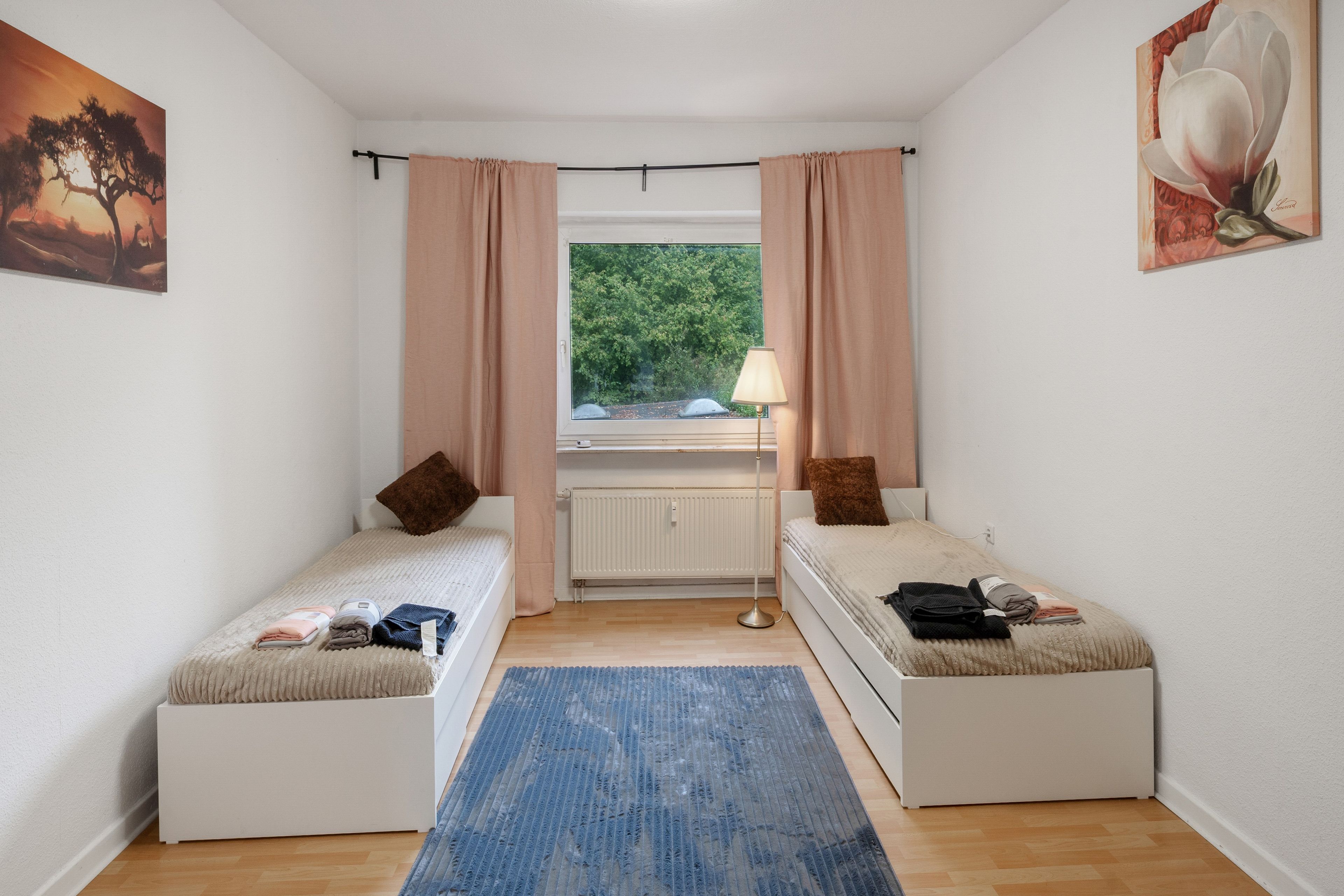 Ein kleines, helles Schlafzimmer mit zwei Einzelbetten, einem Heizkörper, einem Fenster mit Vorhängen, einer Lampe und Kunstwerken an den Wänden.