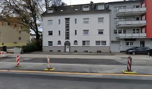 Ein mehrstöckiges Gebäude mit einem zentralen Eingang, Balkonen und einem Parkplatz mit Barrieren.