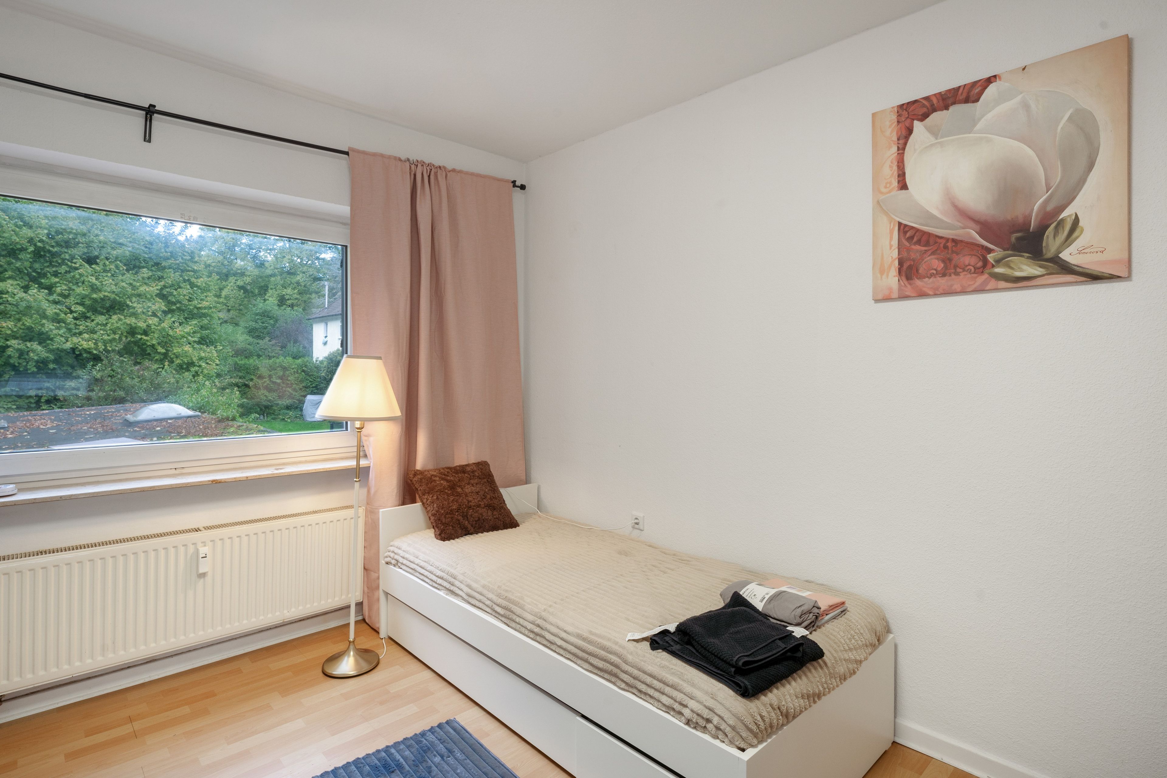 Una habitación pequeña con cama individual, ventana, radiador, lámpara y un cuadro en la pared.