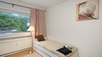 Una habitación pequeña con cama individual, ventana, radiador, lámpara y un cuadro en la pared.
