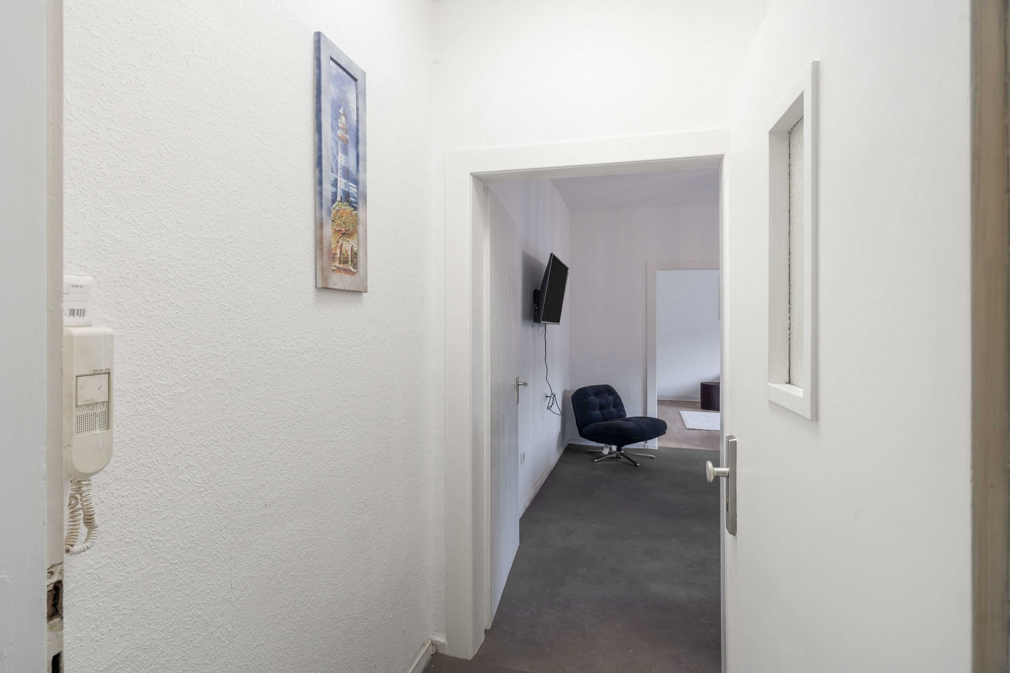 Un pasillo estrecho con pared blanca, una puerta al final, un televisor montado y una silla.