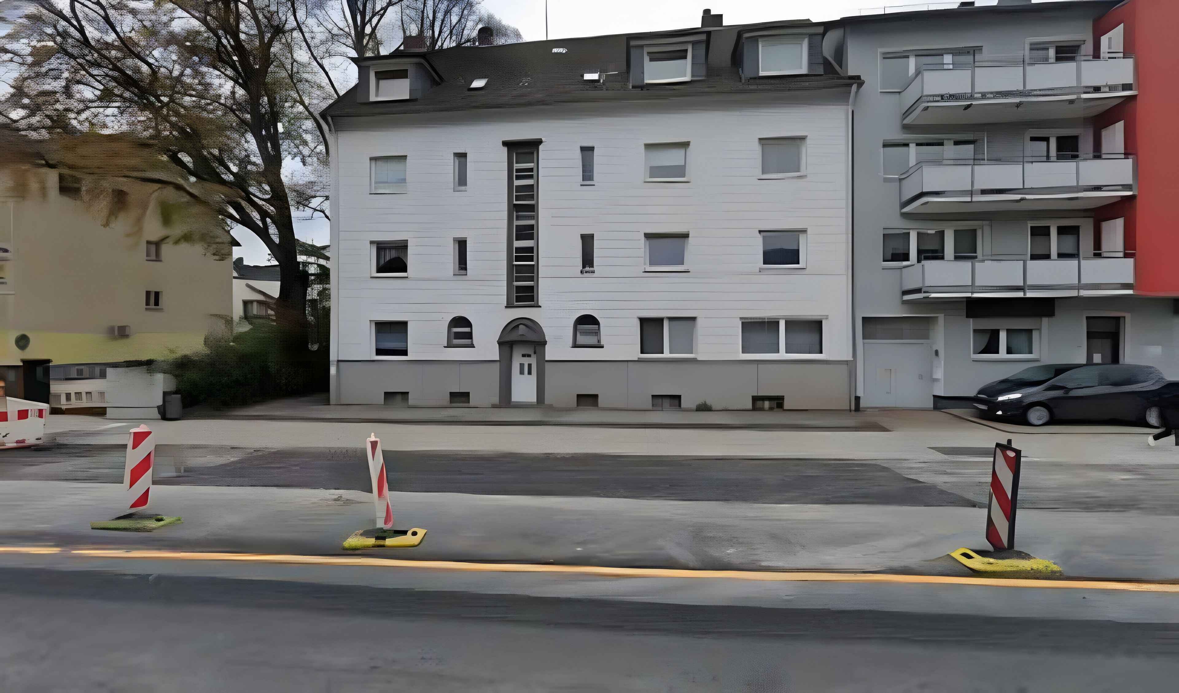 Ein mehrstöckiges Gebäude mit einem zentralen Eingang, Balkonen und einem Parkplatz mit Barrieren.