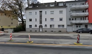 Ein mehrstöckiges Gebäude mit einem zentralen Eingang, Balkonen und einem Parkplatz mit Barrieren.