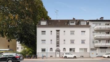 Ein mehrstöckiges Gebäude mit einem zentralen Eingang, einem Parkplatz mit Autos und einem Baum im Vordergrund.