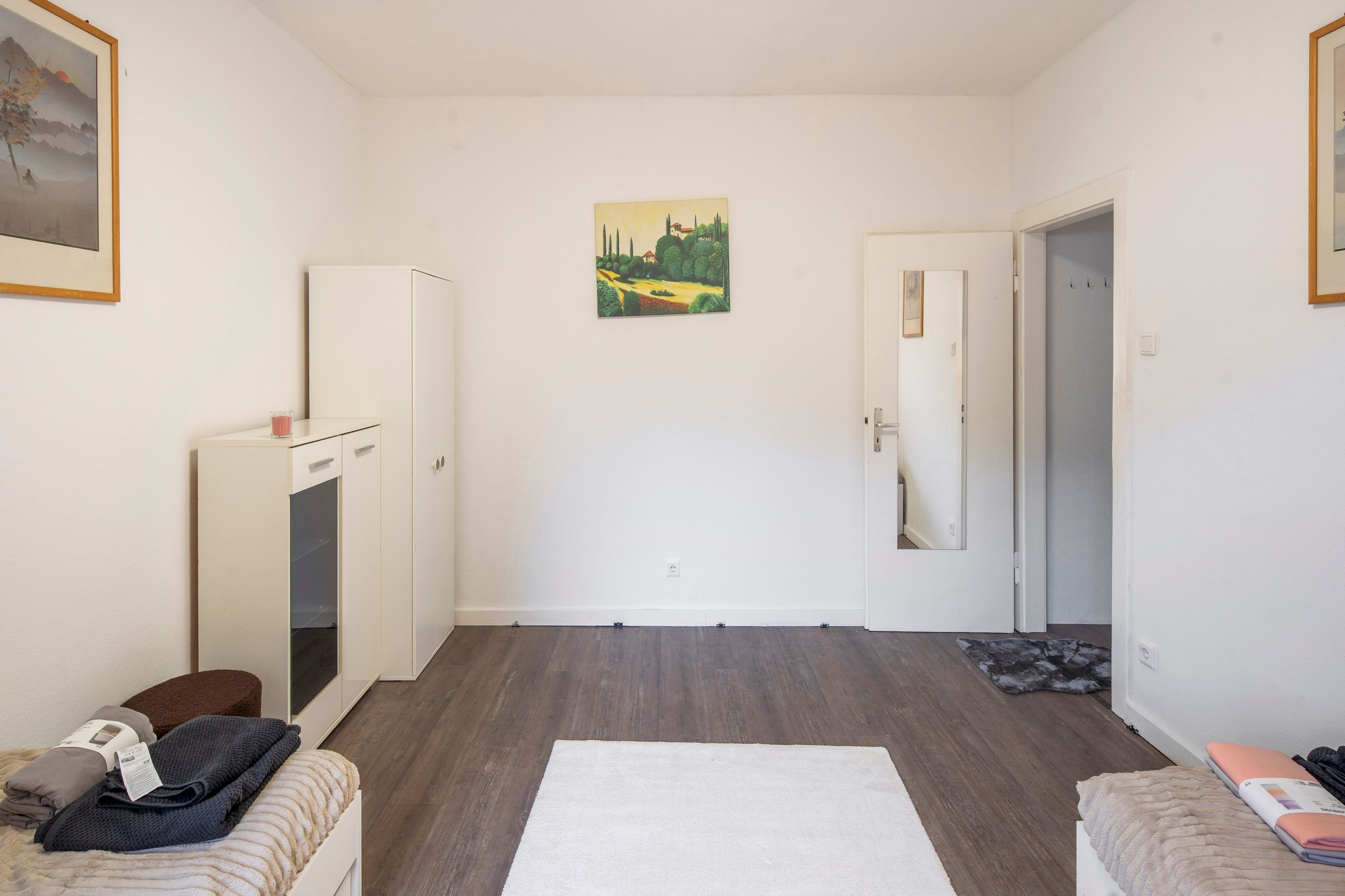 Una habitación con una puerta blanca, un armario blanco, una mesita con un vaso, una alfombra blanca y un cuadro en la pared.