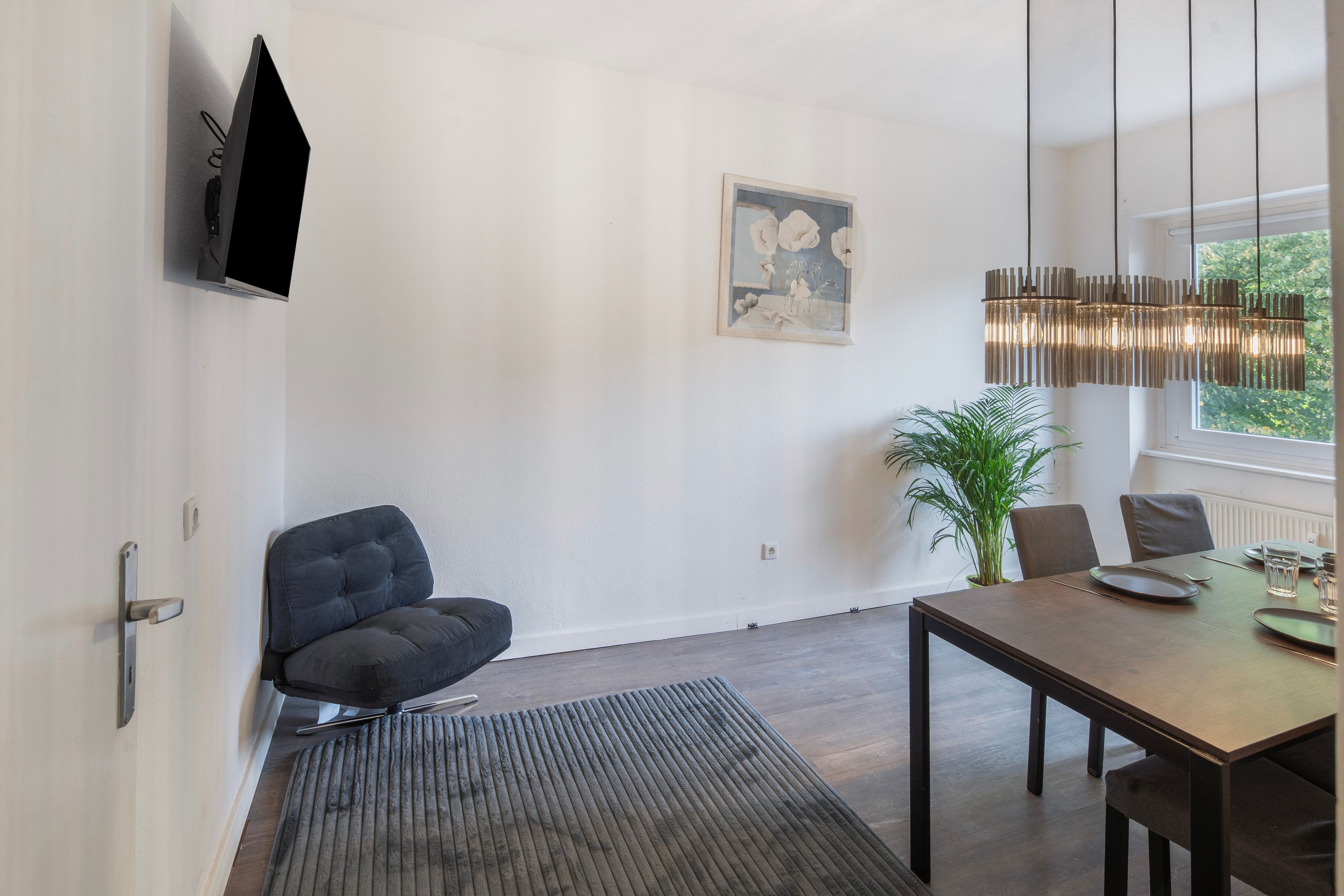 Un salón moderno con un sofá oscuro, una mesa de comedor con sillas, un ventanal con vistas a la vegetación, un televisor montado en la pared y una obra de arte enmarcada.