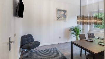 Un salón moderno con un sofá oscuro, una mesa de comedor con sillas, un ventanal con vistas a la vegetación, un televisor montado en la pared y una obra de arte enmarcada.