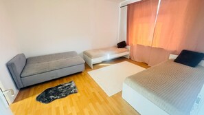 Una habitació amb un sofà gris, un llit amb capçalera blanca, una taula petita blanca i una catifes gran sobre un terra de fusta.