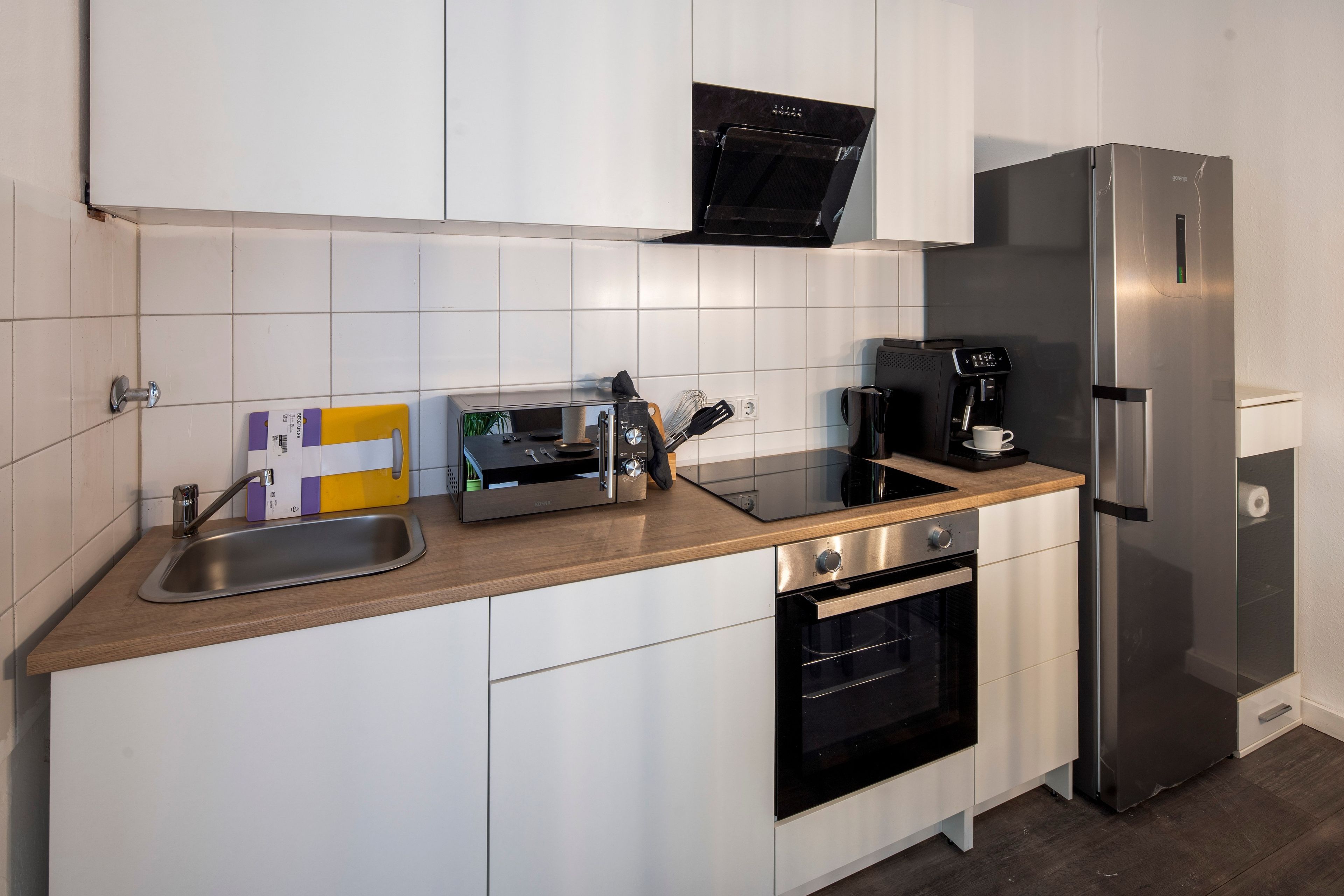 A modern kitchen with white cabinets, a stainless steel refrigerator, a coffee machine, and a built-in oven.