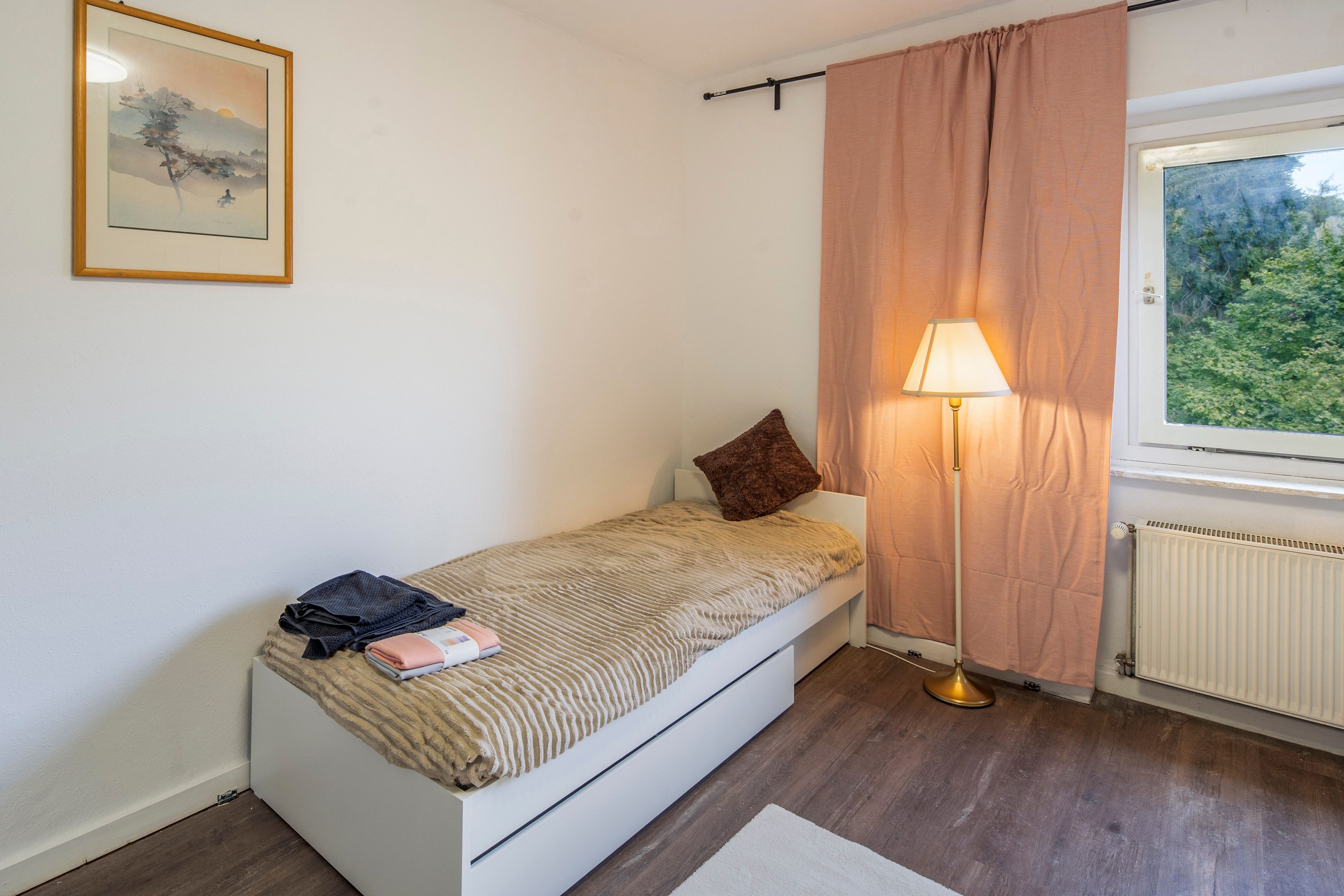 A small, single-bed room with a window, a bedside lamp, and a framed picture on the wall.