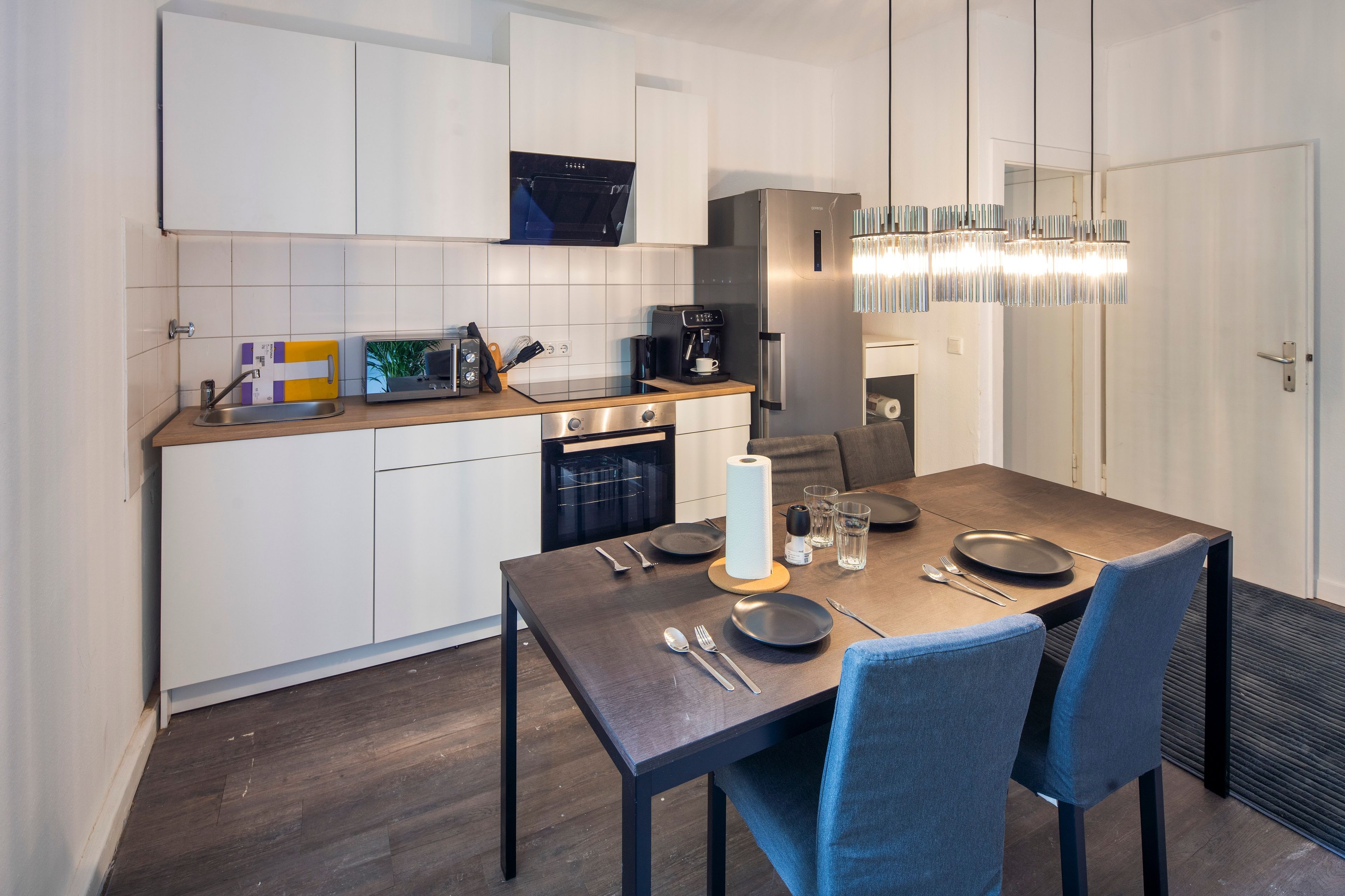 Una cocina moderna con armarios blancos, horno integrado, cafetera y un comedor con una mesa dispuesta para dos.