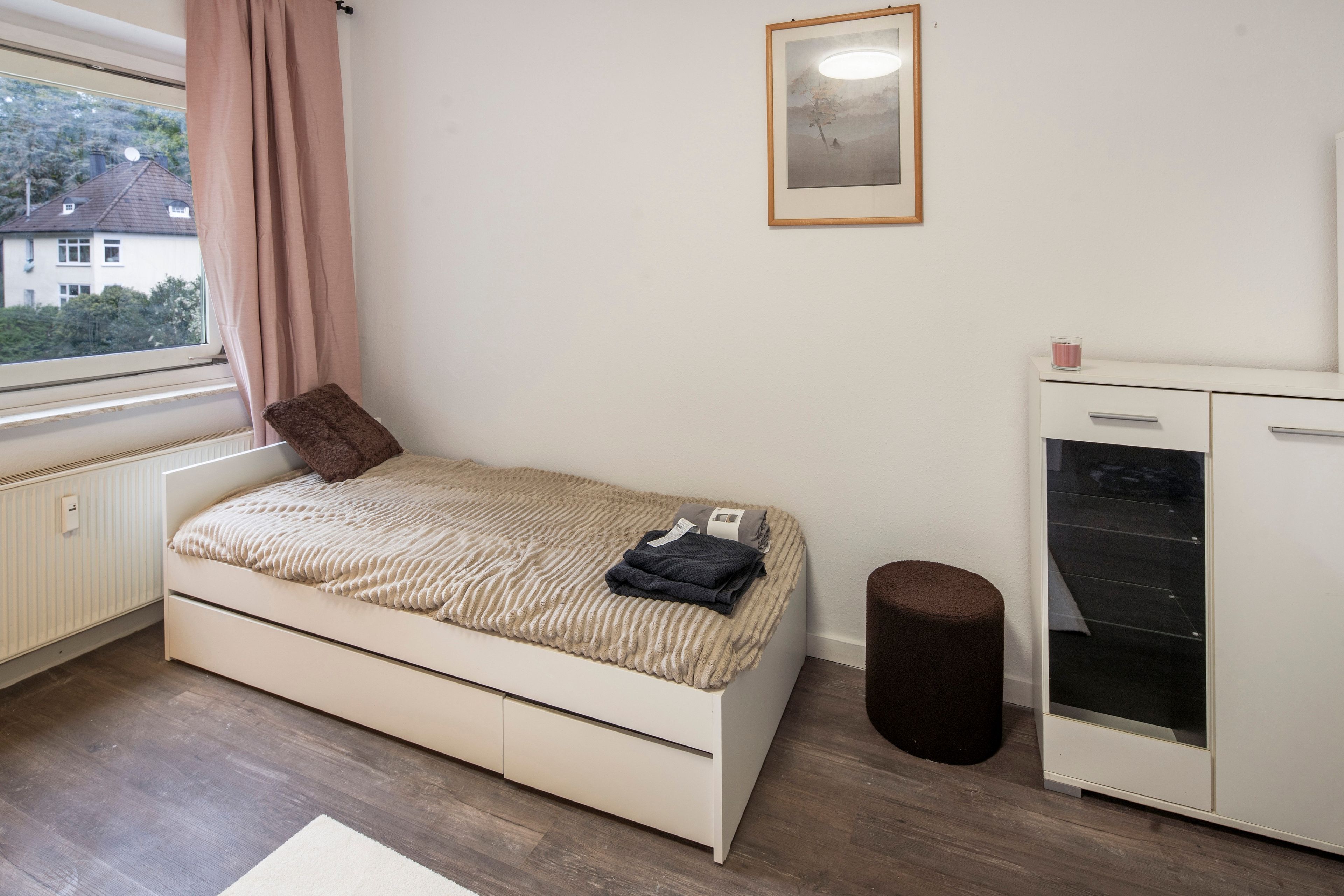 Ein Schlafzimmer mit einem Bett, einem kleinen Hocker, einer Heizung, einem Fenster mit Vorhängen, einem gerahmten Bild an der Wand und einem weißen Schrank.