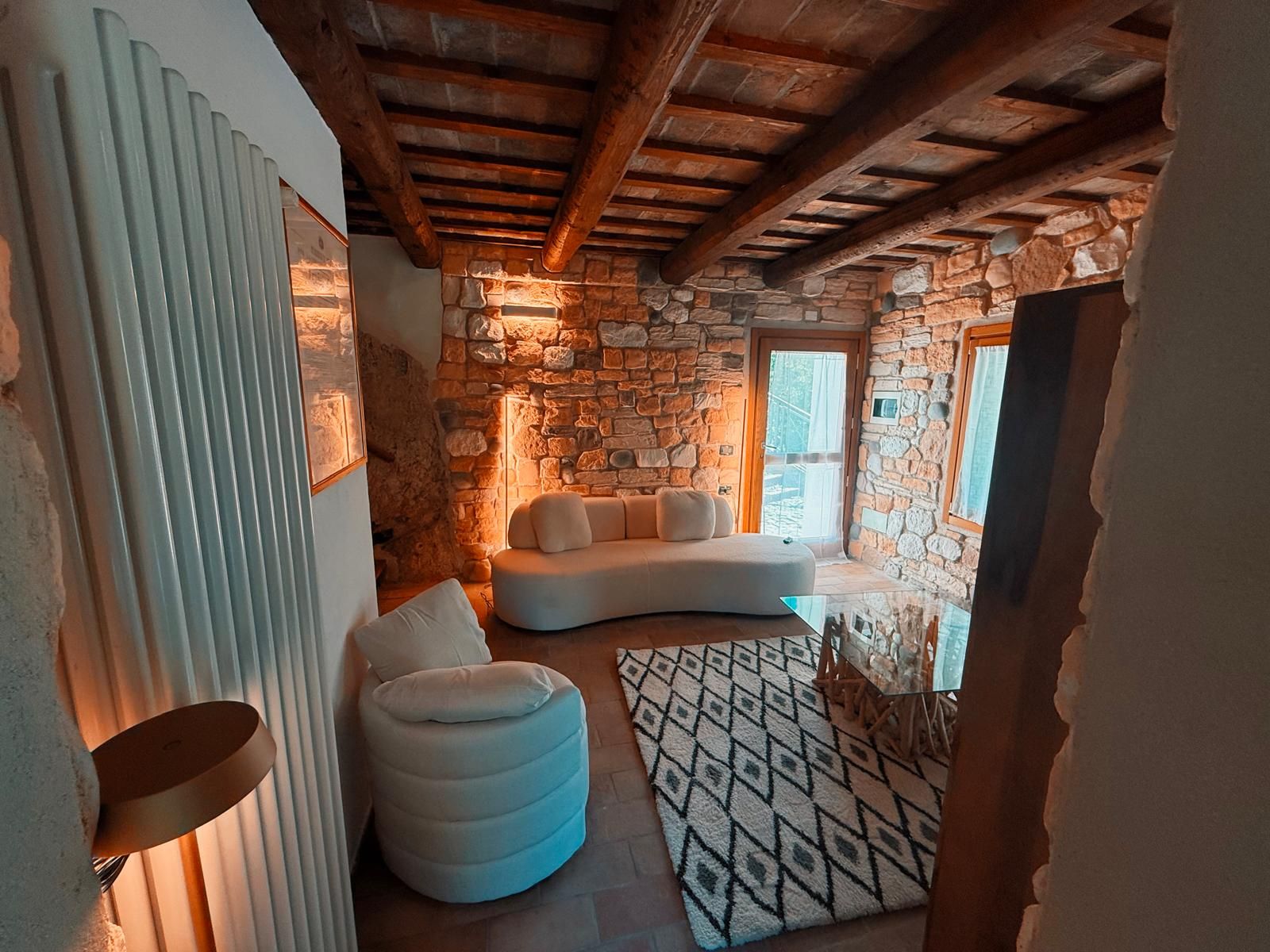 Une pièce chaleureuse avec un mur en pierre, un canapé blanc, un pouf rond et un tapis à motifs.