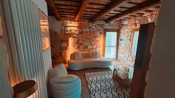 Une pièce chaleureuse avec un mur en pierre, un canapé blanc, un pouf rond et un tapis à motifs.