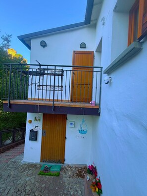 Un edificio de dos pisos con un balcón, una puerta de madera y un pequeño jardín con flores.