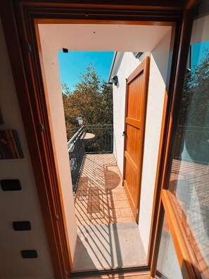 Blick durch eine Glastür auf einen Balkon mit einer Holztür, einem kleinen Tisch und einem Stuhl.