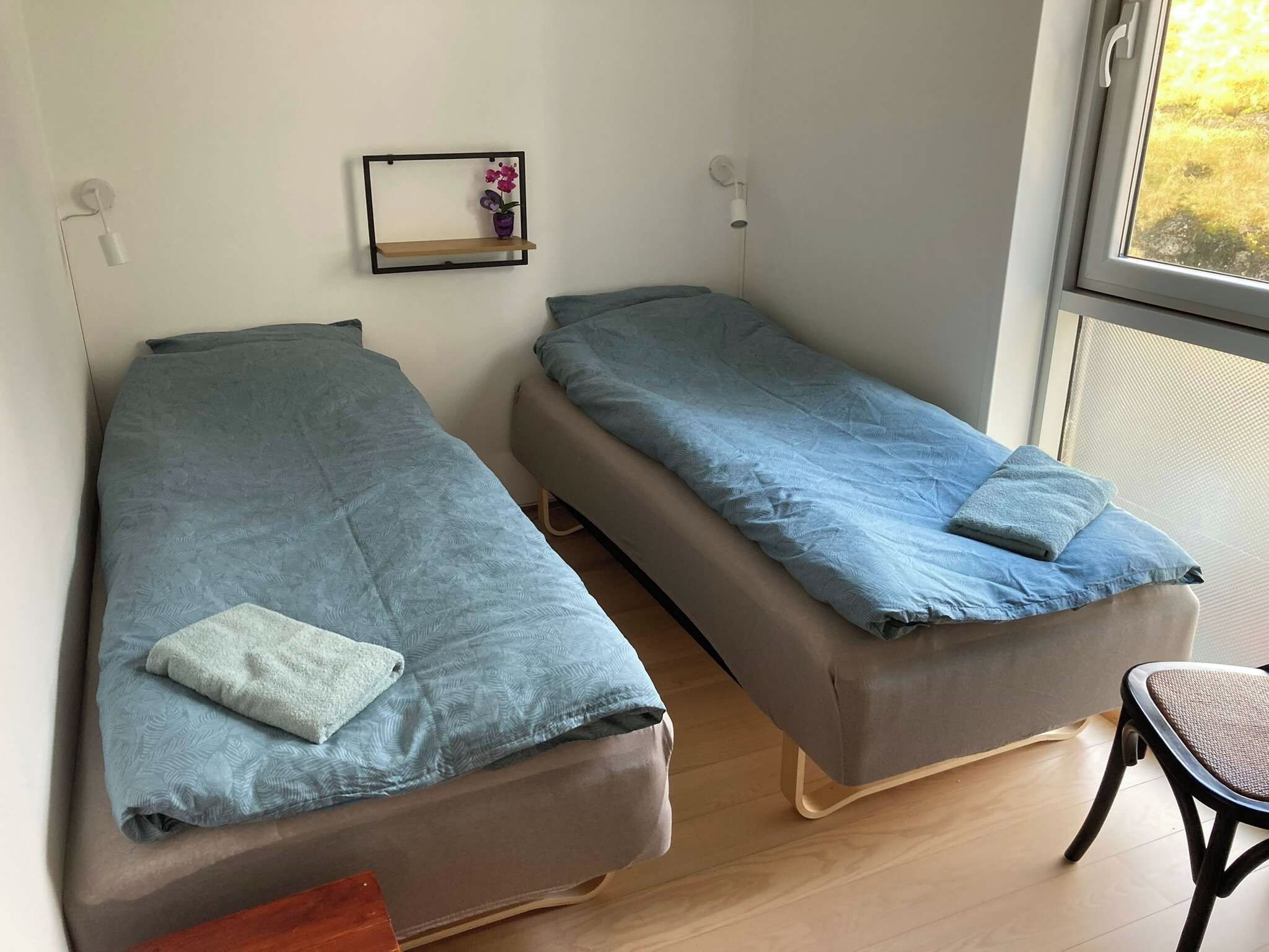 A room with two single beds, a small shelf with a vase, and a chair.
