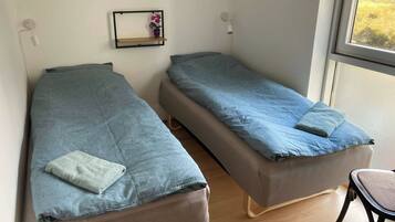 A room with two single beds, a small shelf with a vase, and a chair.