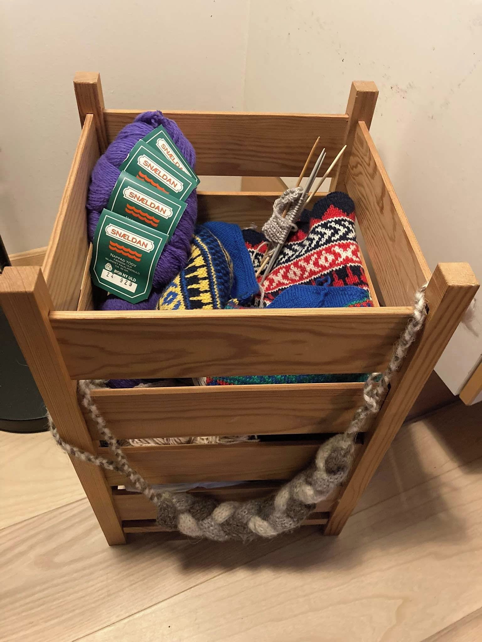A wooden crate with a handle, containing various items including a purple knitted item, a multicolored knitted item, and a green package labeled 'SNELDAN'.