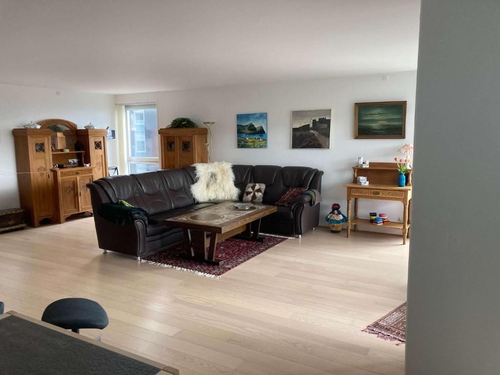 A living room with a sofa, a coffee table, and a dining table. There are paintings on the wall and a cabinet.