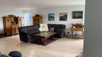 A living room with a sofa, a coffee table, and a dining table. There are paintings on the wall and a cabinet.