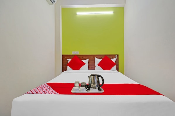 A hotel room with a bed, a bedside table with a kettle, and a red and white bedspread.