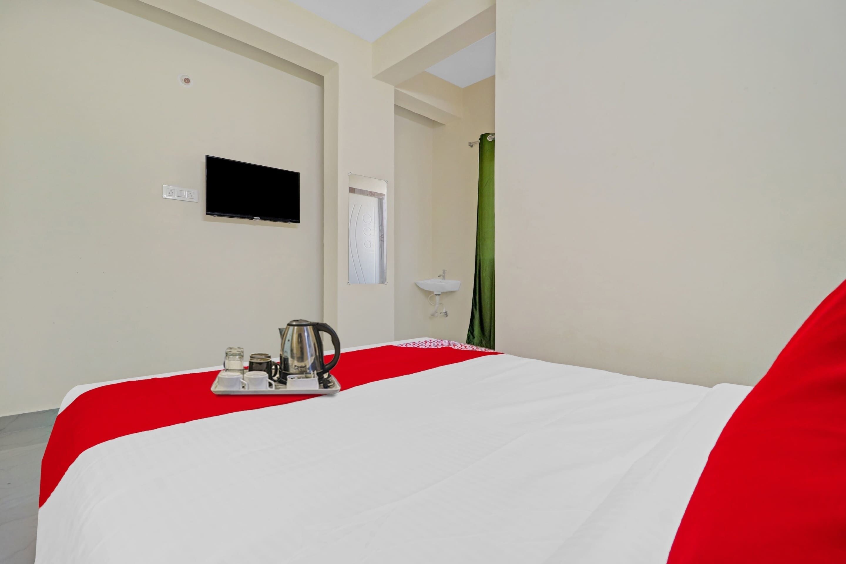 Una habitación de hotel con cama, televisor montado en la pared, una mesita con tetera y tazas, y una cortina verde.