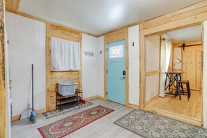 A room with a blue door, a white wall, a wooden floor, a red and beige patterned rug, a metal shelf with a white basket, and a broom. - Lovely 2 bedroom apartment with patio and yard near downtown hospital (Casper)