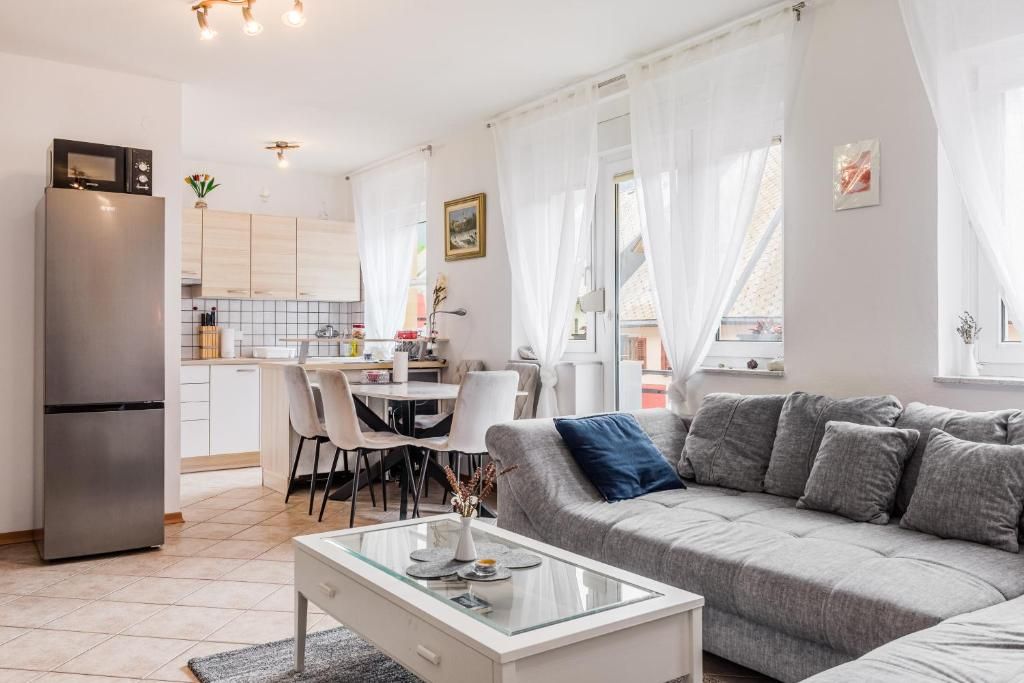 Ein moderner Wohnraum mit grauem Sofa, einem Glastisch und einer Küchenzeile mit weißen Schränken und einem Kühlschrank.