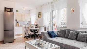 Ein moderner Wohnraum mit grauem Sofa, einem Glastisch und einer Küchenzeile mit weißen Schränken und einem Kühlschrank.