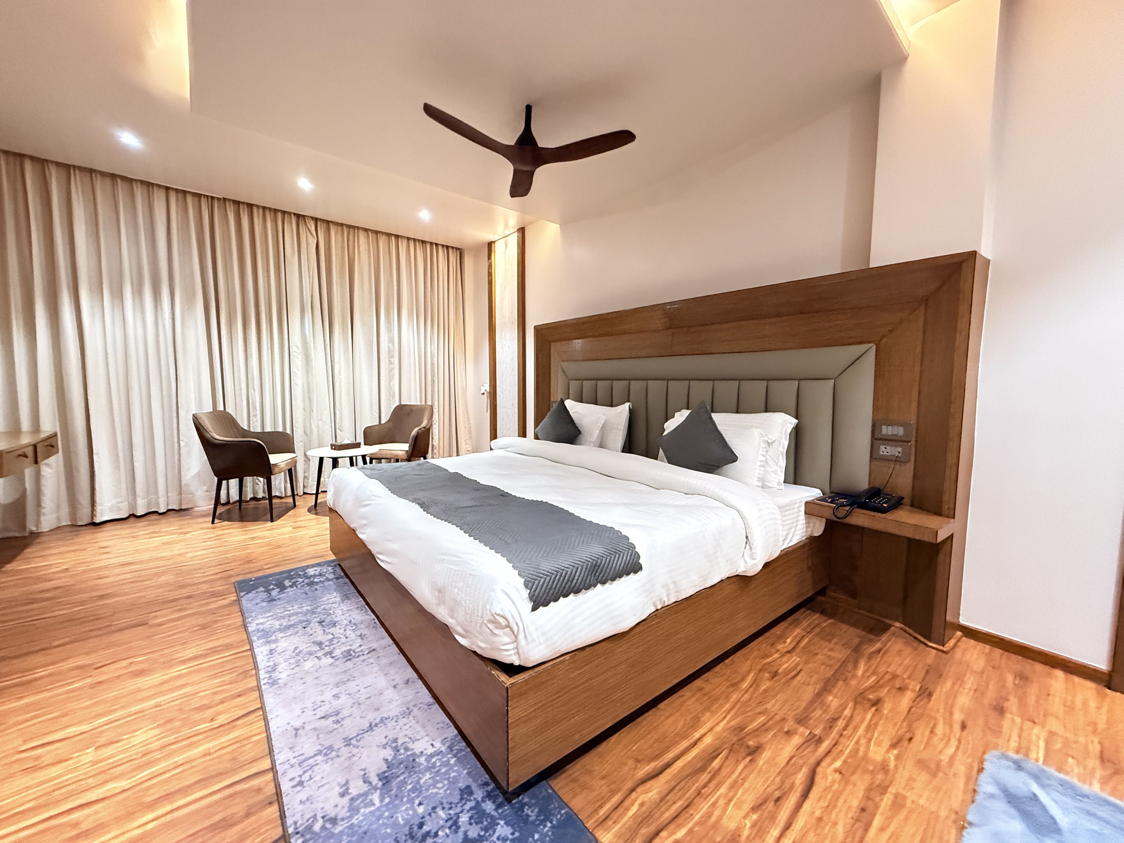 Habitación de hotel moderna con una cama grande, cabecero de madera, un escritorio, dos sillas y un ventilador de techo.