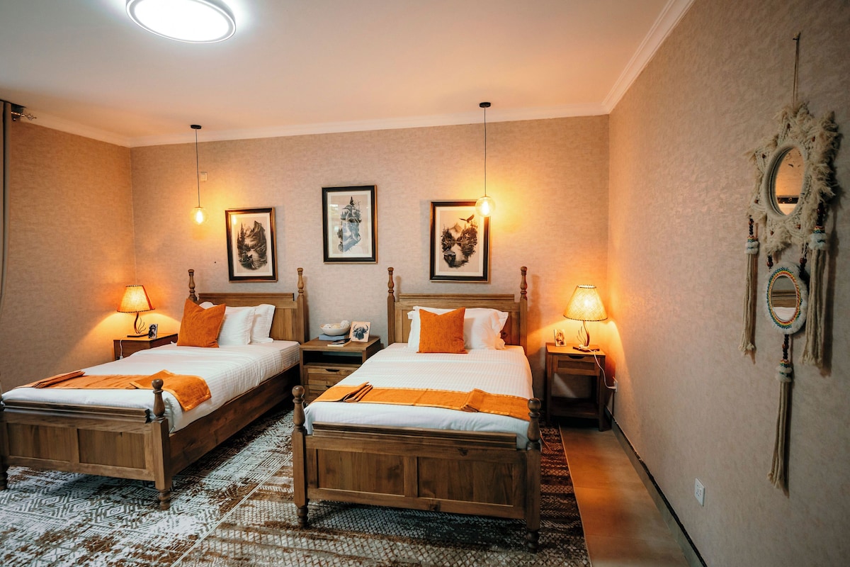 A hotel room with two wooden beds, a patterned rug, and framed artwork on the walls.