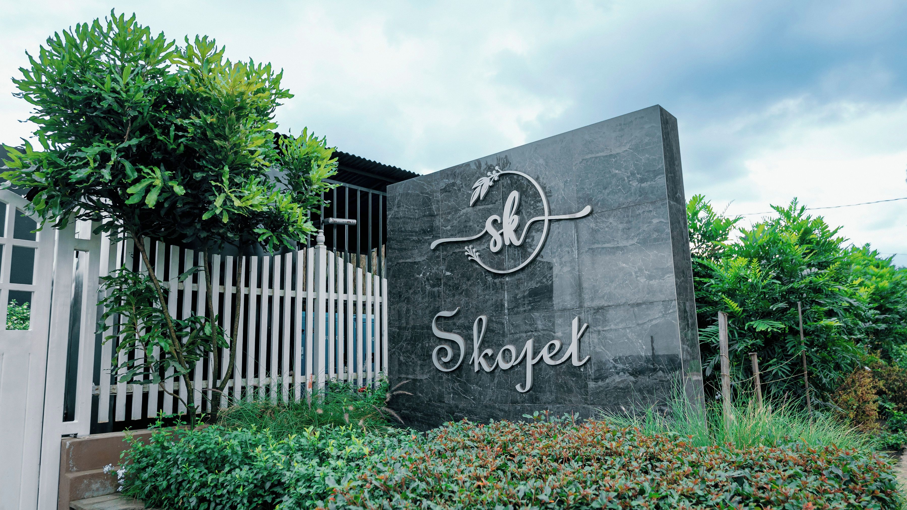 A sign with the name 'Skogel' on a dark stone wall, surrounded by greenery and a white fence.
