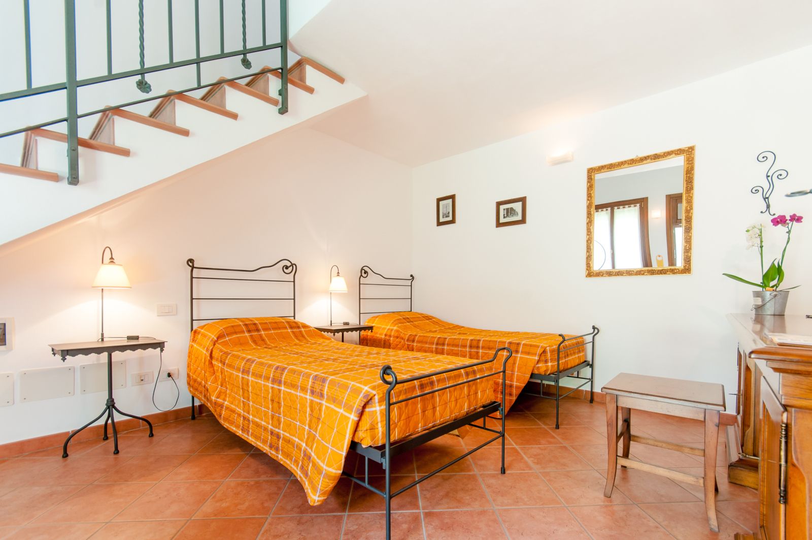 A bedroom with two beds, a staircase, a bedside table with a lamp, a mirror, and a small desk with a chair.
