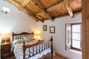 A bedroom with a wooden ceiling, a bed with floral bedding, a nightstand, a window with curtains, and wall-mounted lights. - Casa del Guarda - Finca & Spa Vereda Real (Albacete)