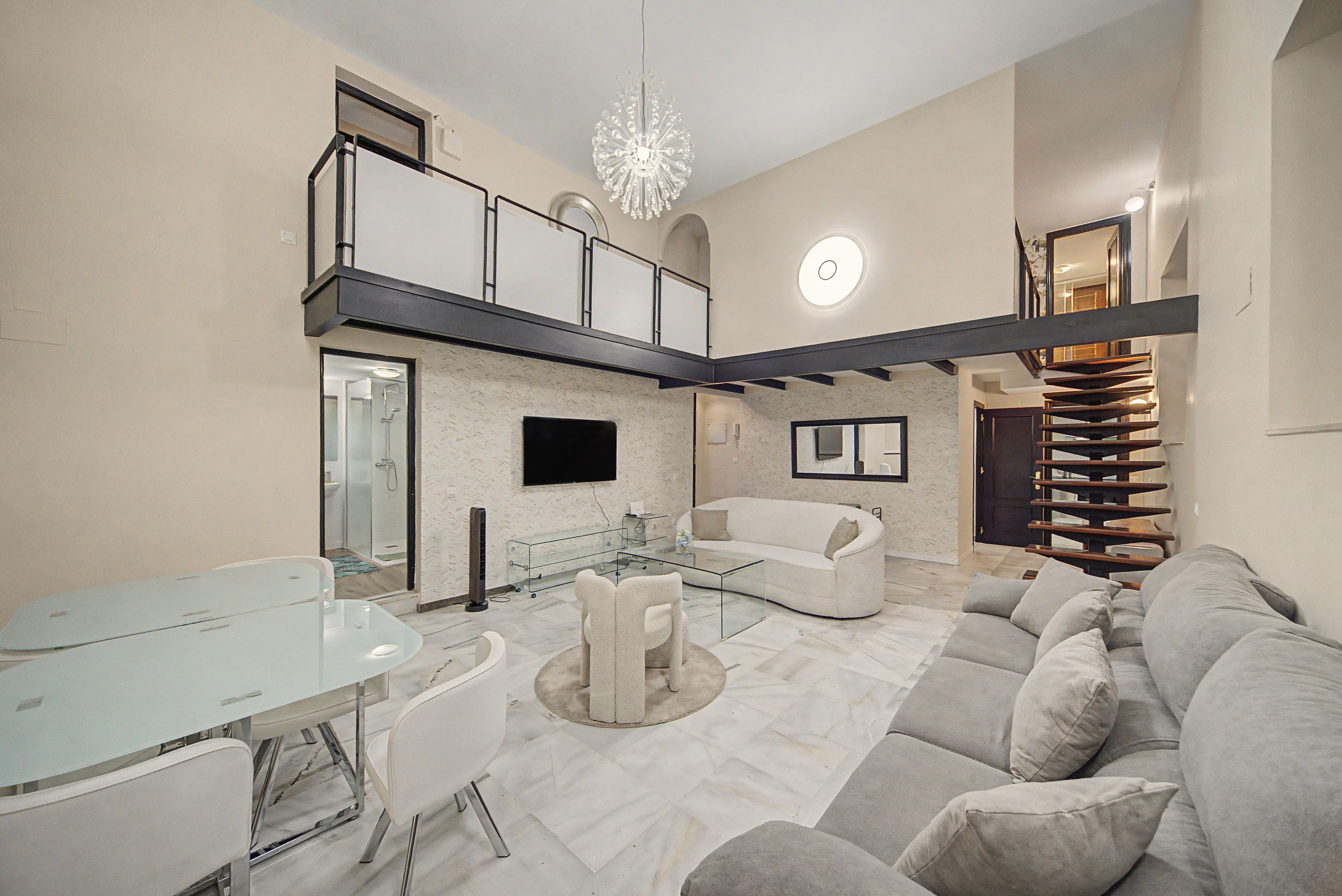 A modern living room with a glass dining table, a sofa, and a TV mounted on the wall.