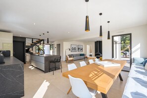 A modern kitchen with a central island, a dining area with a wooden table, and a living room with a sofa and a fireplace. - Vrbo Property (Pyla-sur-Mer)