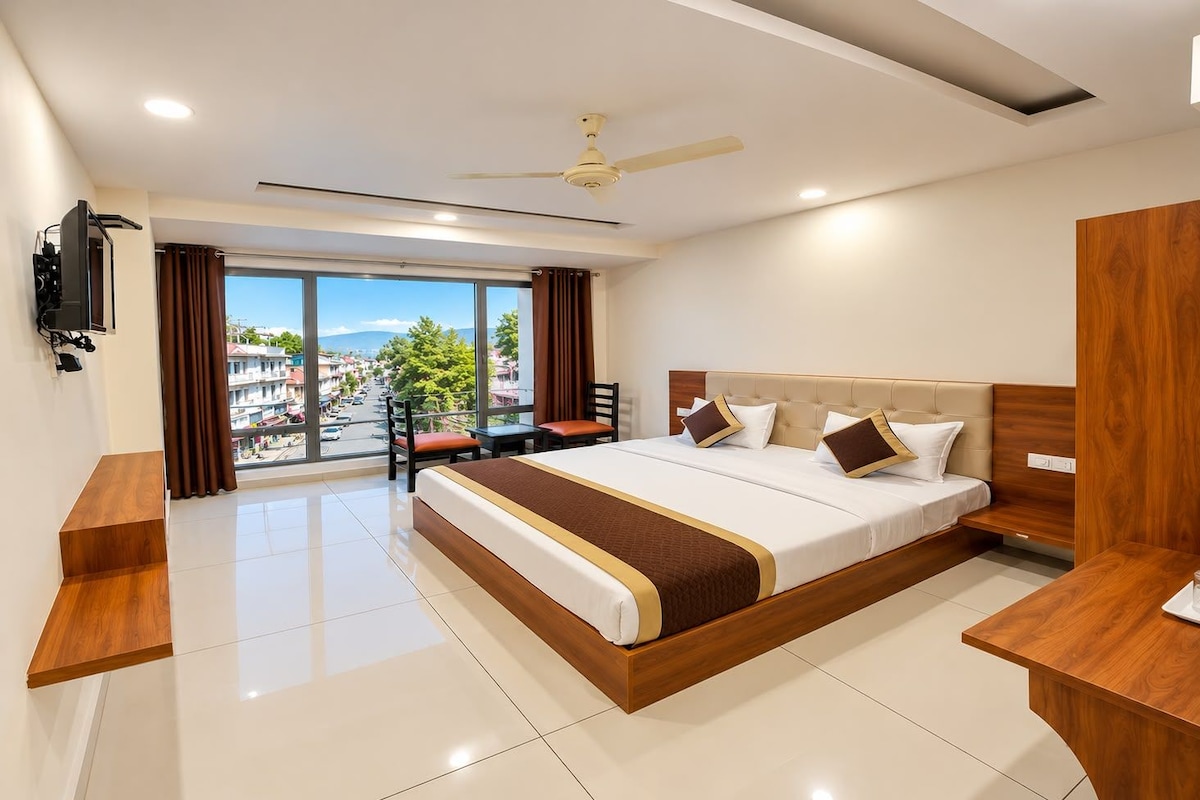 Habitación de hotel con una cama grande, un escritorio, una silla, un televisor y vistas de la ciudad a través de amplios ventanales.
