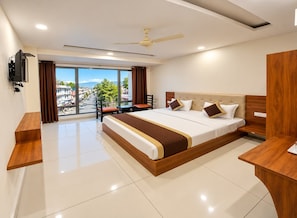 Habitación de hotel con una cama grande, un escritorio, una silla, un televisor y vistas de la ciudad a través de amplios ventanales.