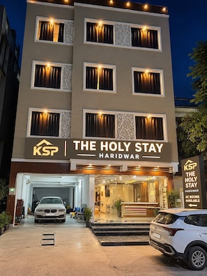 Un hotel llamado 'The Holy Stay' con una fachada moderna, múltiples ventanas con cortinas y un letrero que indica que hay habitaciones con aire acondicionado.