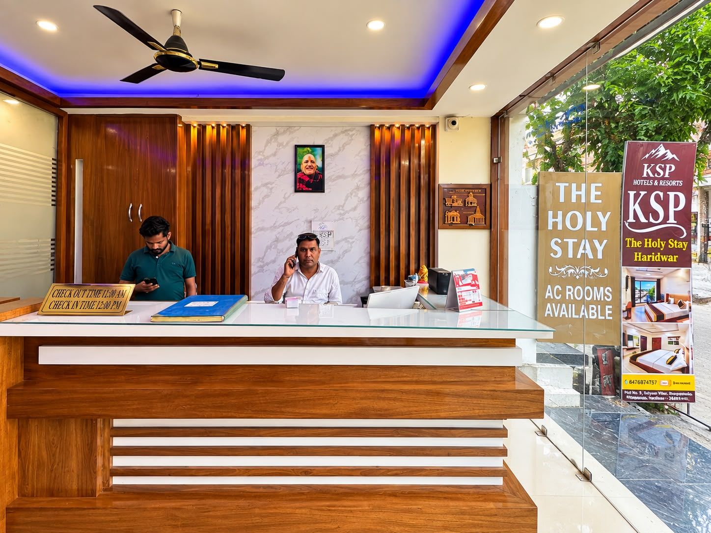 Un comptoir d’accueil d’hôtel avec un guichet d’enregistrement, un ventilateur de plafond et un panneau publicitaire pour les chambres climatisées.