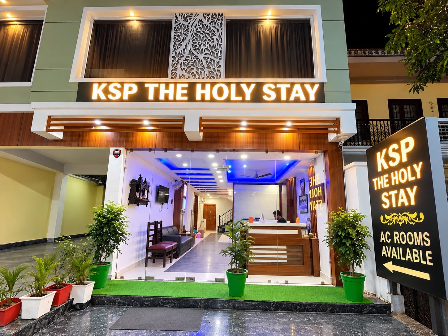 En hotellentrén med en reception, rum med luftkonditionering finns tillgängliga, och en skylt visar hotellets namn: ”KSP THE HOLY STAY”.